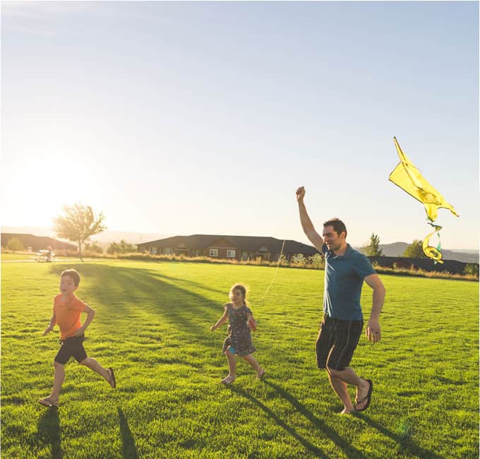 A dad and two kids fly a kite on green grass