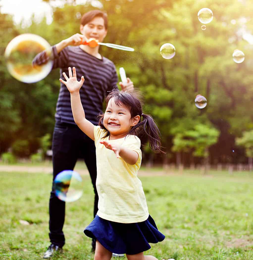 Little girl running after bubbles, dad in background