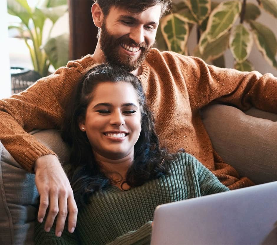 A couple smile while looking at a computer on a couch