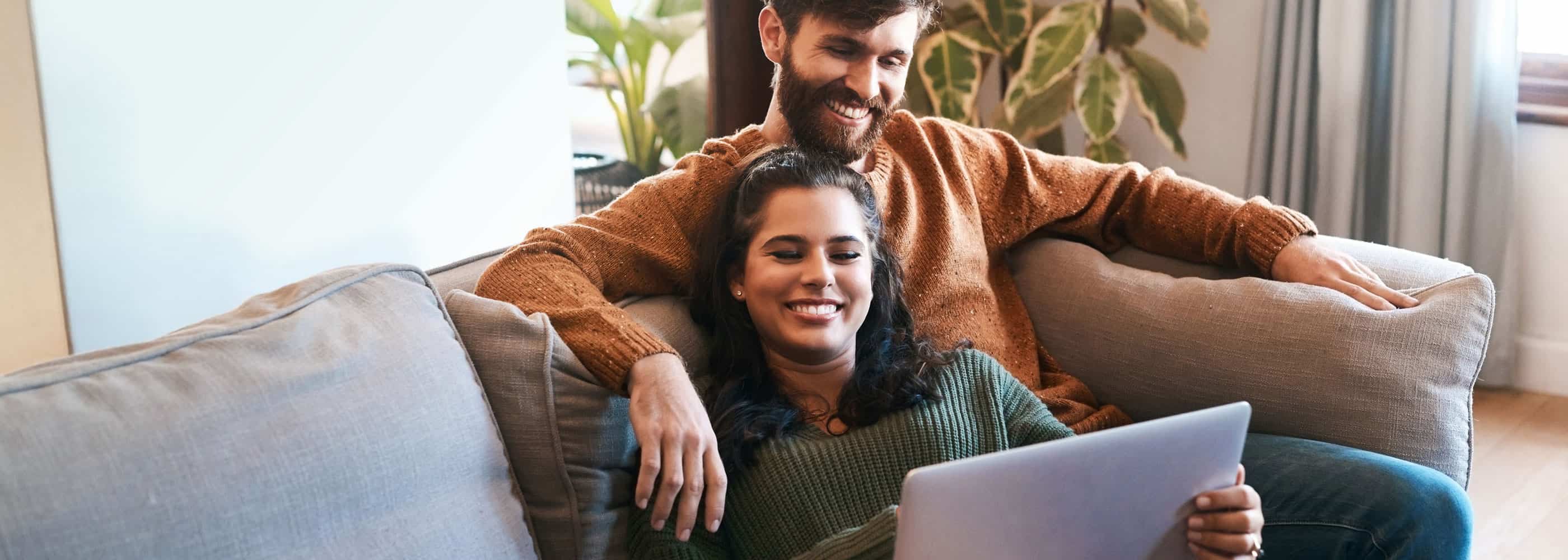 A couple smile while looking at a computer on a couch