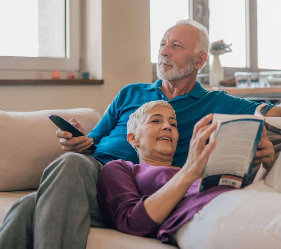 Older couple relax on the couch, man with remote, woman reading a book